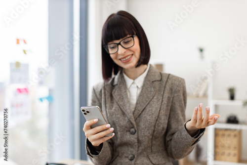 Thoughtful Caucasian professional talking during online presentation at staff meeting. Beautiful smiling woman in suit standing near office table having video call.
