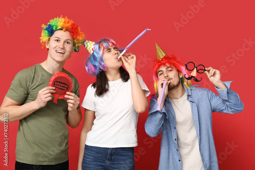 Young friends in funny disguise with part whistles and whoopee cushion on red background. April fool's day celebration photo