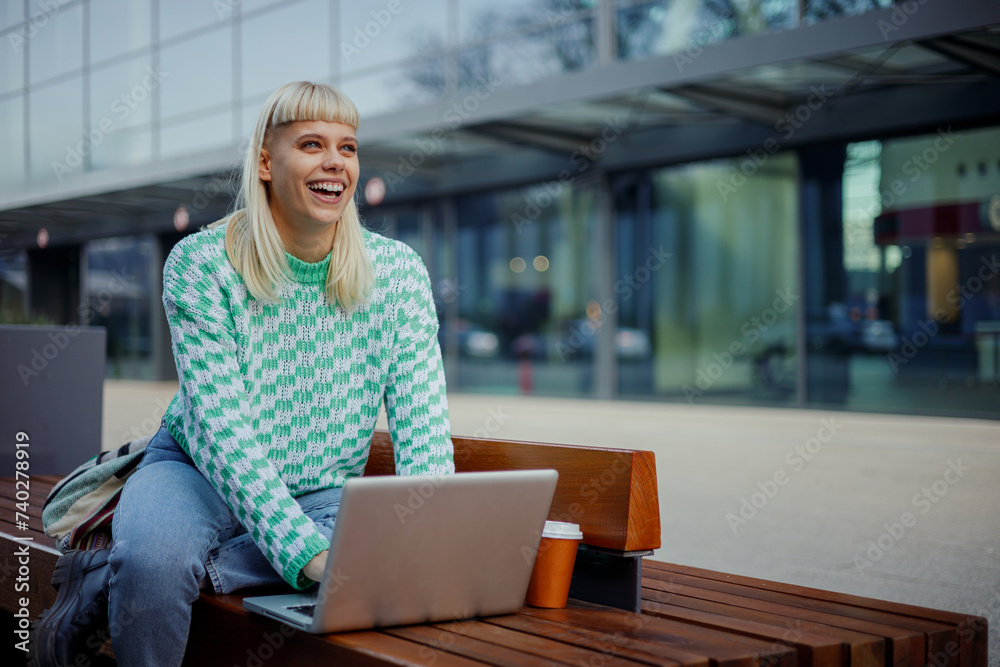 Obraz premium A happy college girl is sitting at campus and typing on a laptop.