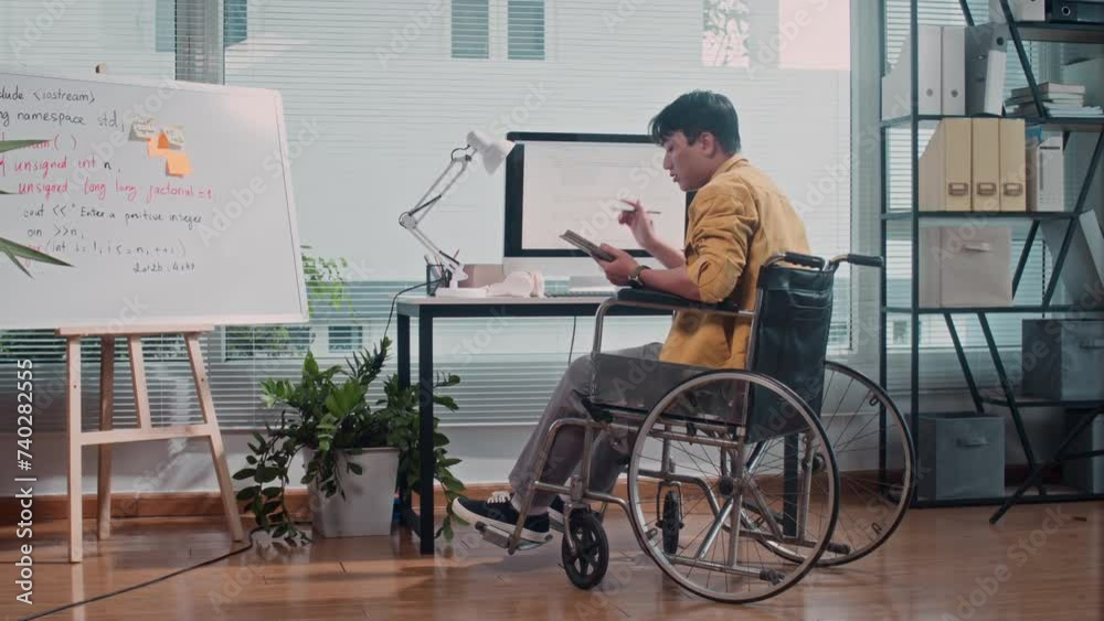 Long shot, back view of pensive male coder in wheelchair working on computer at office desk Long shot, back view of pensive male coder in wheelchair working on computer at office desk