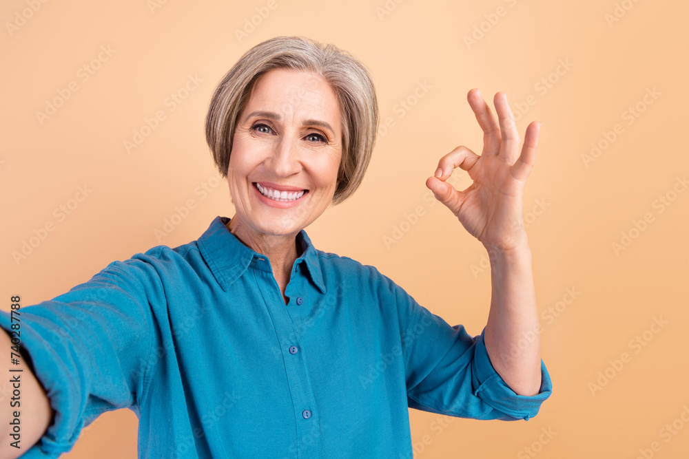 Photo of toothy beaming woman with bob hairstyle dressed blue shirt making selfie showing okey symbol isolated on beige color background
