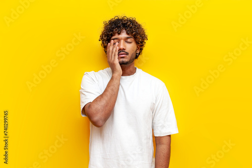 tired indian man in white t-shirt suffering from headache and sad in depression on yellow isolated background, unhappy curly guy rubbing his eyes