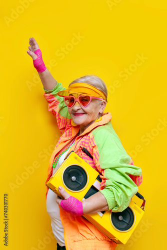 funny granny in hipster clothes listening to music on a tape recorder and showing a kiss on a yellow isolated background, elderly cool woman dancing at a party and having fun