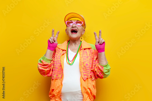 crazy old granny in sports colorful clothes wins and showing peace gesture on yellow isolated background, elderly woman in youth hipster clothes rejoices in victory and luck and screams