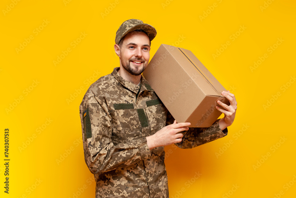 Ukrainian army soldier in pixel camouflage uniform holds a box on a yellow isolated background, Ukrainian military cadet carries and gives humanitarian aid and donation - obrazy, fototapety, plakaty 
