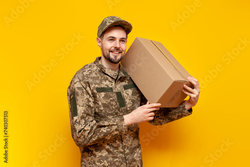 Ukrainian army soldier in pixel camouflage uniform holds a box on a yellow isolated background, Ukrainian military cadet carries and gives humanitarian aid and donation photo