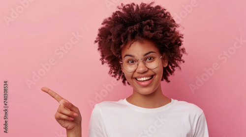 An exuberant young woman with curly hair and glasses, pointing her finger to the side with a wide, joyful smile on a pink background.