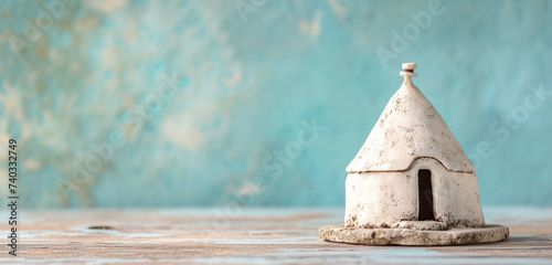 A miniature Italian trullo with a conical roof, on a polished wooden surface. The background is a soft powder blue.