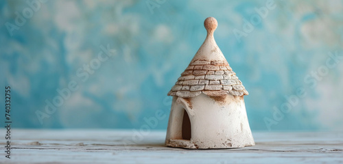 A miniature Italian trullo with a conical roof, on a polished wooden surface. The background is a soft powder blue.