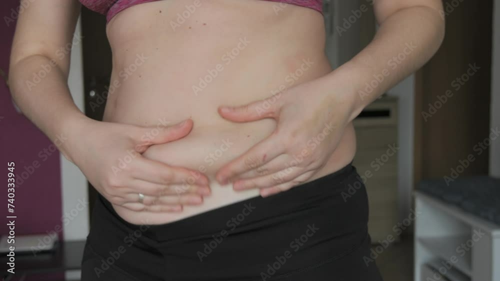 Female belly body positive. Woman touches belly folds in flat slow motion. A small layer of fat on abdomen of woman wearing a sports bra and leggings. Concept of sport, healthy lifestyle or overweight Female belly body positive. Woman touches belly folds in flat slow motion. A small layer of fat on abdomen of woman wearing a sports bra and leggings. Concept of sport, healthy lifestyle or overweight