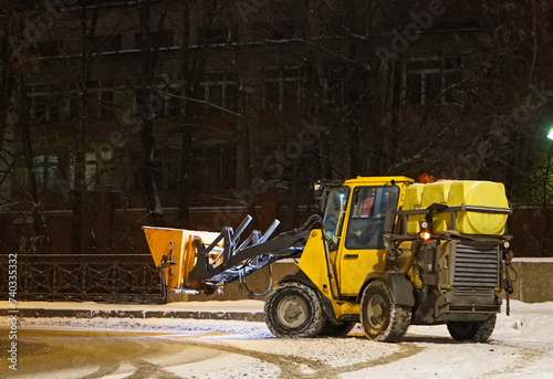 A snowplow cleans the sidewalks. A snowplow clears the street of snow at night. Selective focus. Russian winter. Snow removal.	
