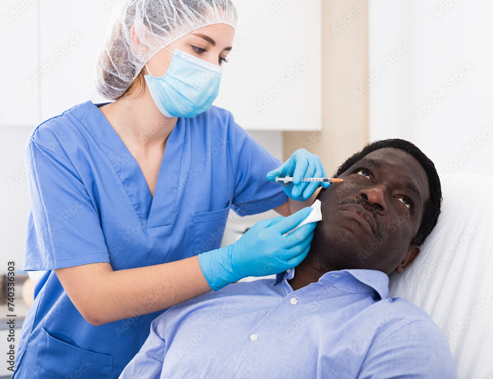 Fototapeta premium Skilled female cosmetologist performing face contouring injections to adult african american man in cosmetology clinic. Male skin care concept