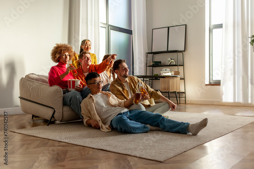 multiracial group of young friends sitting on the couch at home drinking beer and watching TV