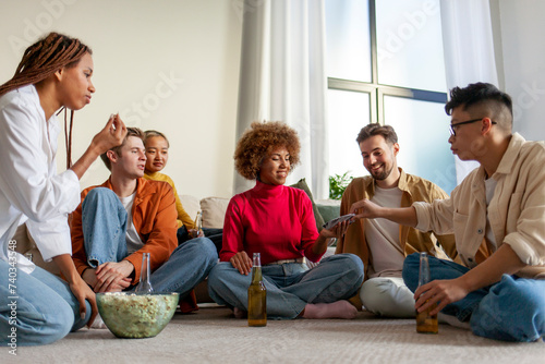 multiracial group of young people sitting at home with beer and popcorn and playing cards with friends