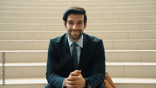 Smiling skilled businessman looking at camera while sitting at stairs. Young project manager smile at camera while clasping hands at outdoor with blurred yellow background. Front view. Exultant. photo