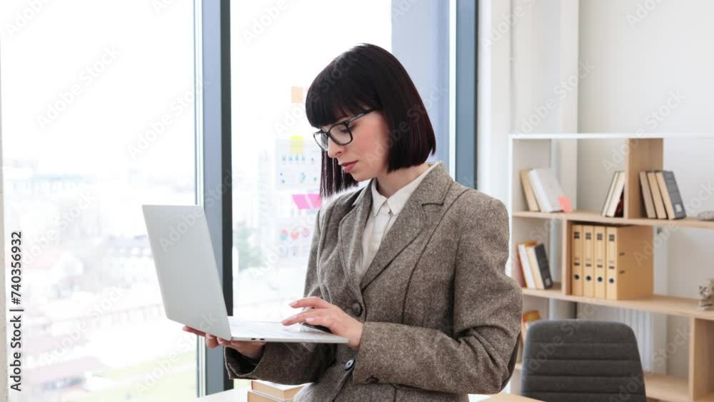 Beautiful smiling woman in suit standing with portable computer near office table. Thoughtful caucasian professional preparing for online presentation during staff meeting. Beautiful smiling woman in suit standing with portable computer near office table. Thoughtful caucasian professional preparing for online presentation during staff meeting.