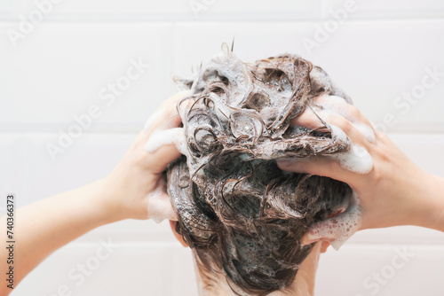 Fototapeta Naklejka Na Ścianę i Meble -  A young woman washes her hair with shampoo on white tiles background, back view.