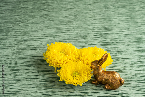 Decorative gold Easter Bunny sniffing three large yellow chrysanthemum flowers on a textured green velvet background, Happy Spring
