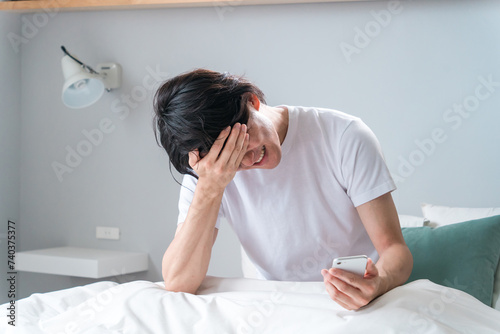 ベッドの上でスマホを見てショックを受けるアジア人男性（寝坊・遅刻・失恋・炎上・大暴落）
 photo