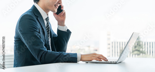 オフィスでノートパソコンを使いながら電話する若いアジア人ビジネスマン
