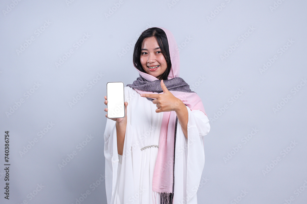 Young Asian muslim woman wearing hijab showing and pointing finger to blank phone screen for mockup Young Asian muslim woman wearing hijab showing and pointing finger to blank phone screen for mockup
