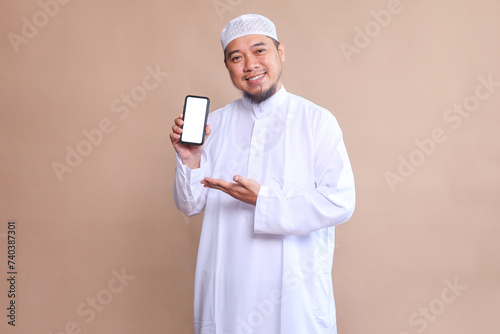 Asian muslim man in white robe showing phone screen over beige background