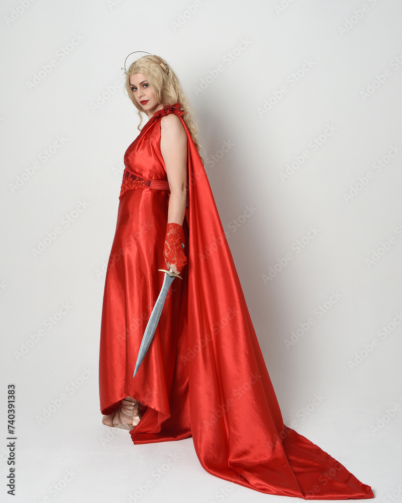 Full length portrait of beautiful blonde model dressed as ancient mythological fantasy goddess in flowing red silk toga gown, crown. walking pose, holding a sword weapon, isolated studio background Full length portrait of beautiful blonde model dressed as ancient mythological fantasy goddess in flowing red silk toga gown, crown. walking pose, holding a sword weapon, isolated studio background