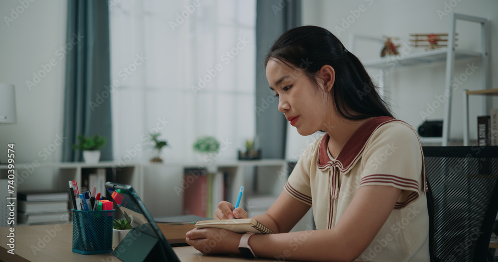 Selective focus, Side view, Female writer sitting at desk thinking idea holding pen making notes in diary while working on tablet for online learning at home, creative thoughts to journaling Selective focus, Side view, Female writer sitting at desk thinking idea holding pen making notes in diary while working on tablet for online learning at home, creative thoughts to journaling