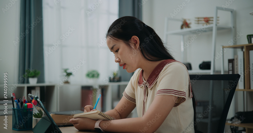 Selective focus, Side view, Female writer sitting at desk thinking idea holding pen making notes in diary while working on tablet for online learning at home, creative thoughts to journaling