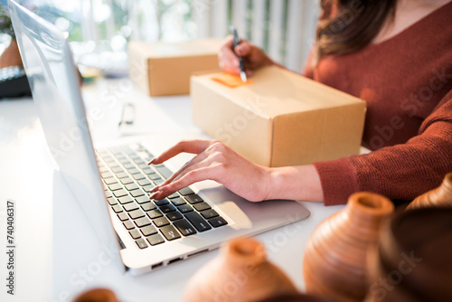 Asian beautiful woman sell vase products online live streaming at home. A young attractive girl writes the address of the customer on the parcel to send. Remote buying and purchase shopping concept.