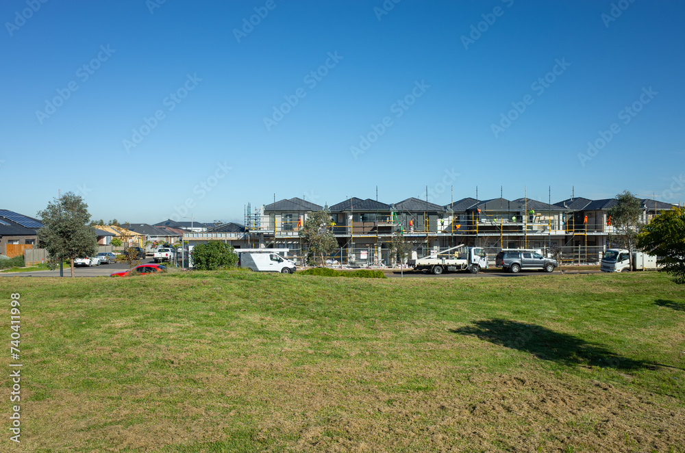 A vacant lot of land with grass lawn with some new townhouses under construction in the background. Concept of development in a new residential neighbourhood, suburban homes and Australia real estate. A vacant lot of land with grass lawn with some new townhouses under construction in the background. Concept of development in a new residential neighbourhood, suburban homes and Australia real estate.
