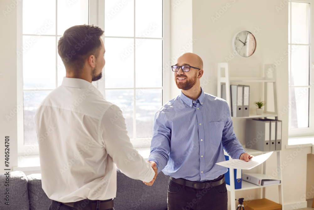 Fototapeta premium Two happy smiling men shaking hands standing in office finishing meeting, celebrating success, business achievement, making a good deal, , signing a contract or greeting new employee.