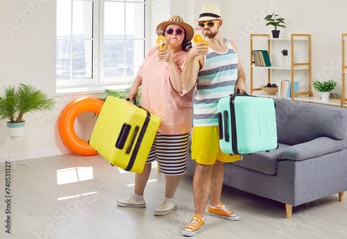 Happy couple of fat woman and smiling man in hats and sunglasses getting ready for holiday trip standing at home with suitcase, beach ball and rubber ring. Vacation and summer journey concept.