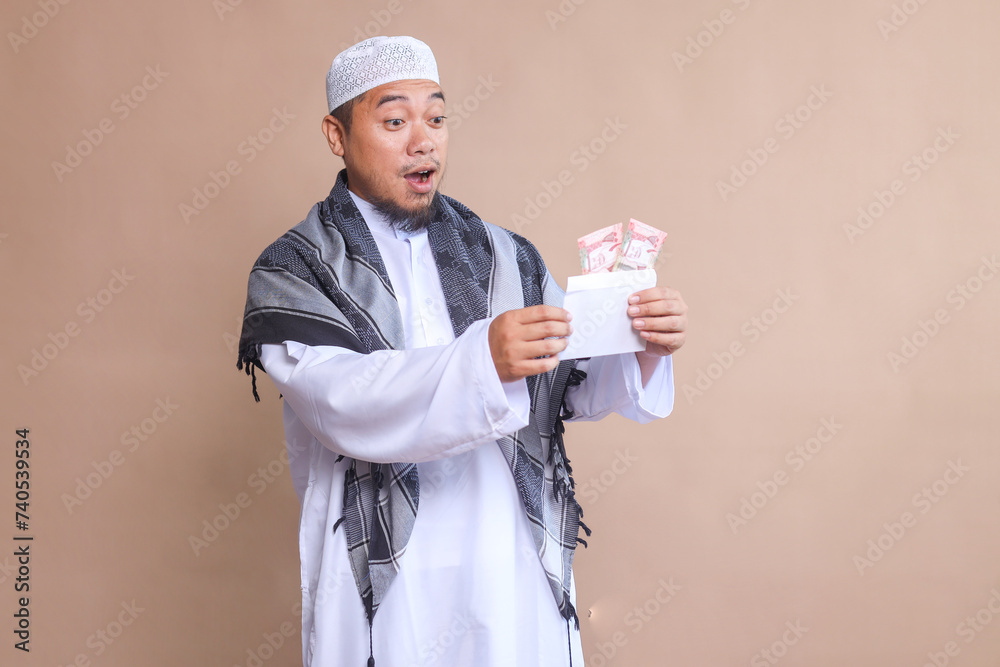 Surprised Muslim Asian man holding envelope filled with money on beige background Surprised Muslim Asian man holding envelope filled with money on beige background
