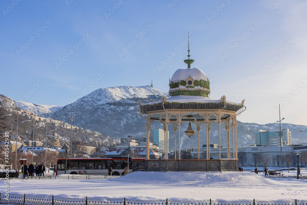 Musical pavilion in central Bergen, covered in snow and in cold but sunny weather. People walking past, magical bergen backdrop in the background.