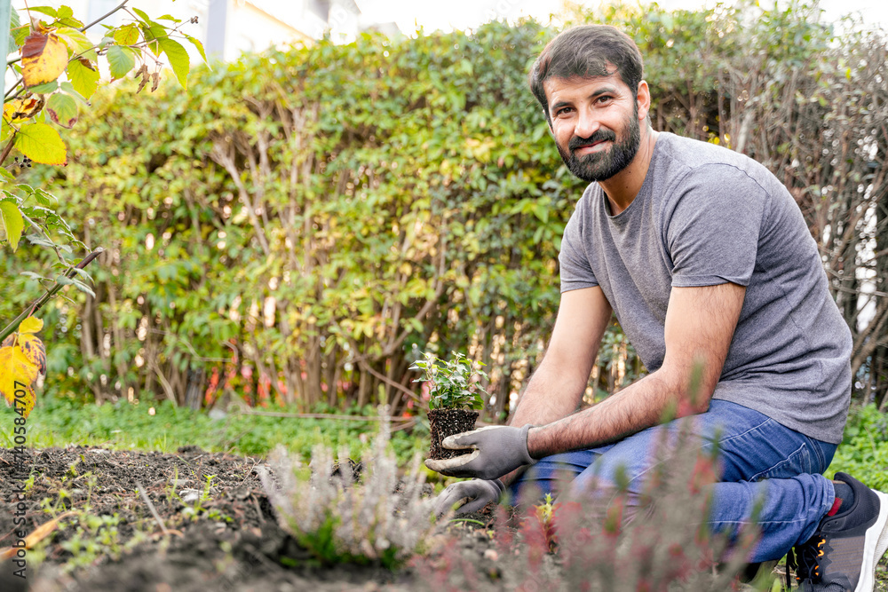 Pakistani or indian man with a beard, wearing jeans, is kneeling in a garden holding a potted plant. He is surrounded by grass, trees, shrubs, and has a smile on his face Pakistani or indian man with a beard, wearing jeans, is kneeling in a garden holding a potted plant. He is surrounded by grass, trees, shrubs, and has a smile on his face