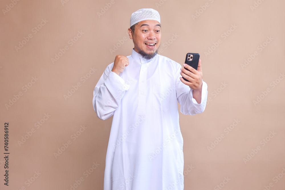 Asian Muslim man clenched fist showing excitement when looking to mobile phone Asian Muslim man clenched fist showing excitement when looking to mobile phone