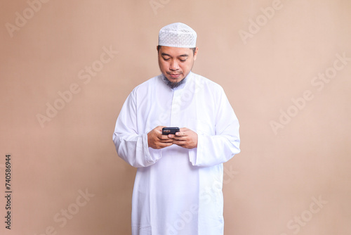 Religious Asian man in white robe and skullcap using mobile phone over beige background photo
