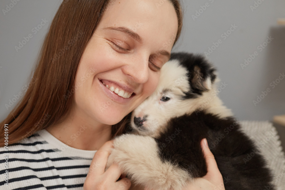 Closeup of smiling cheerful brown haired woman dressed in striped shirt embracing small puppy while sitting on sofa at home in living room expressing happiness