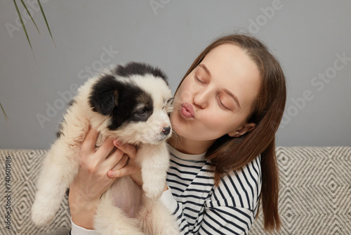 Brown haired woman dressed in striped shirt embracing small puppy while sitting on sofa at home in living room keeps lips pout kissing her charming dog
