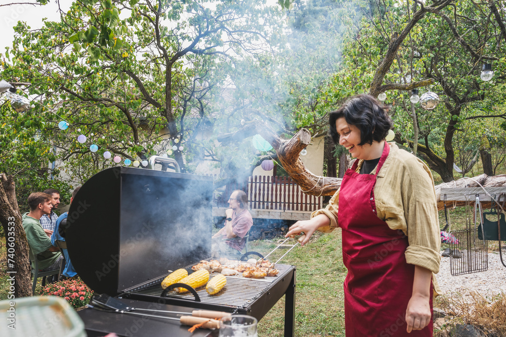 Young happy woman in apron grilling barbecue in garden for friends at country house party
