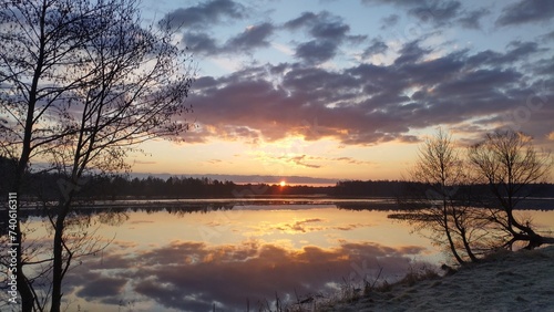 The sun rises from behind the forest on the far side of the lake and colors the sky with bright colors Trees and sky with clouds are reflected in the water The grass on the shore is covered with frost