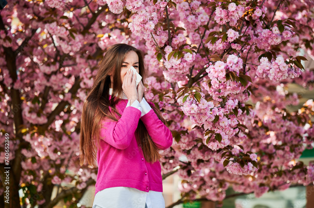 Woman allergic suffering from seasonal allergy at spring in blossoming garden at springtime. Woman sneezing and blowing nose using nasal handkerchief in front of blooming tree. Spring allergy concept. Woman allergic suffering from seasonal allergy at spring in blossoming garden at springtime. Woman sneezing and blowing nose using nasal handkerchief in front of blooming tree. Spring allergy concept.