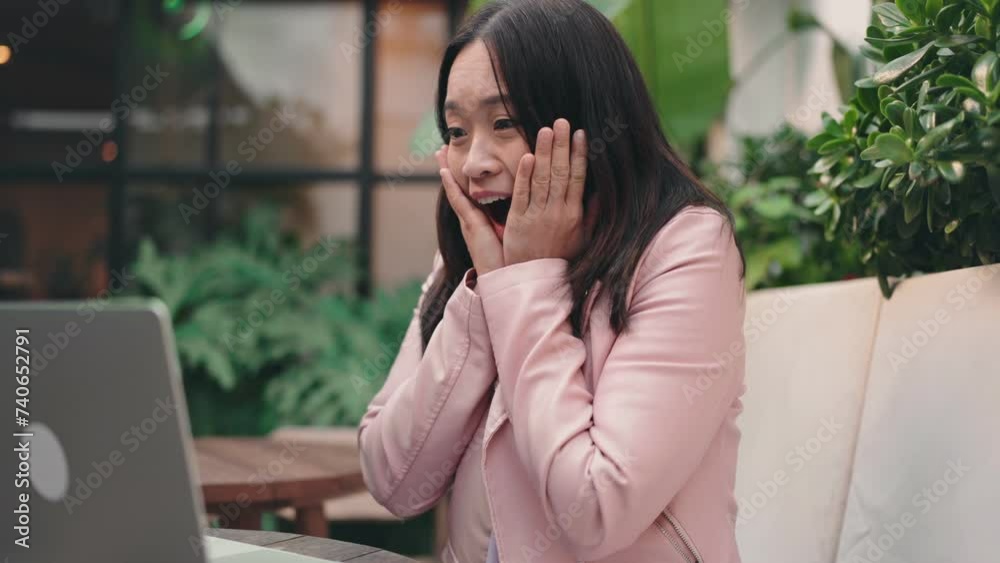 Shocked young Asian woman expressing excitement and happiness while looking at laptop screen in a cozy cafe setting. Shocked young Asian woman expressing excitement and happiness while looking at laptop screen in a cozy cafe setting.