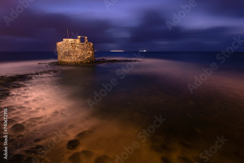 Twilight envelops an ancient stone fortress standing in shallow waters off the coast of Gran Canaria, under a dramatic evening sky photo