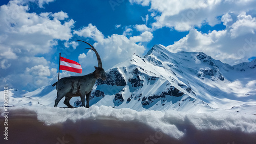 Statue of wooden mountain goat with panoramic view of snow covered mountain peaks of High Tauern along Grossglockner high alpine road, Carinthia Salzburg, Austria. Austrian flag waving in the wind