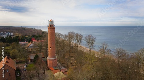 A drone captures Gąski beach, West Pomeranian Voivodeship, Poland, featuring a red brick lighthouse, Baltic Sea, sandy shore, leafless trees on dunes, holiday cottages, hotels, and residences. 