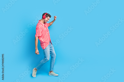 Full body photo of satisfied guy wear stylish shirt jeans trousers in headphones dance near empty space isolated on blue color background