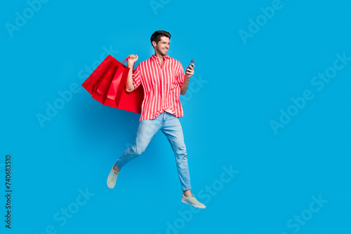 Full body photo of smart guy wear stylish shirt jump hold shopping bags make online order on smartphone isolated on blue color background