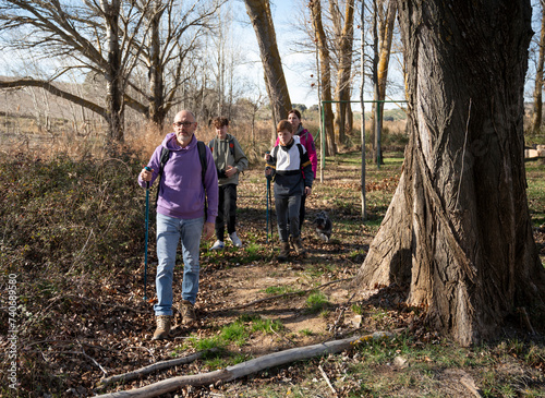 A multi-generational family walking along a forest trail, with a mature man leading the group, followed by young adults and a child, all equipped with trekking poles photo